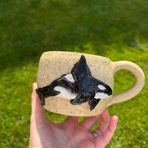May include: A ceramic mug with a speckled cream-colored glaze featuring a raised, glossy black and white orca whale sculpture on the side. The mug has a rounded body and a curved handle, showcasing a textured finish and a detailed marine animal design.