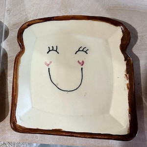 Könnte beinhalten: Keramikplatte in Brotlaibform mit einem lächelnden Gesicht. Die Platte hat eine cremefarbene Mitte mit einem braunen Rand und zeigt schwarze Wimpern, rosa Herzen und einen schwarzen, gebogenen Mund.