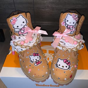 以下が含まれることがあります： ハローキティのワッペン、ピンクのサテンリボン、白いロープの靴紐、ゴールドチェーン、散りばめられたパールとラインストーンで装飾されたタンカラーのレザーブーツ。ブーツのタン部分にもハローキティのワッペンが付いており、箱の上に置かれています。