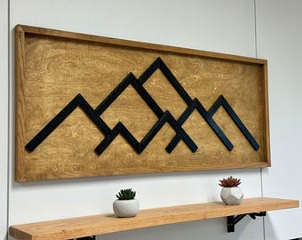 May include: A wooden wall art piece with a black geometric mountain design. The art is framed in a natural wood frame and is mounted on a wall with a shelf below.