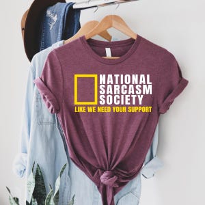 A purple t-shirt with a yellow graphic and text printed on it, hanging on a clothing rack.