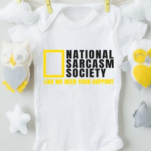 A white baby onesie with the text "NATIONAL SARCASM SOCIETY LIKE WE NEED YOUR SUPPORT" printed on it, along with a yellow square and a heart-shaped plush toy.
