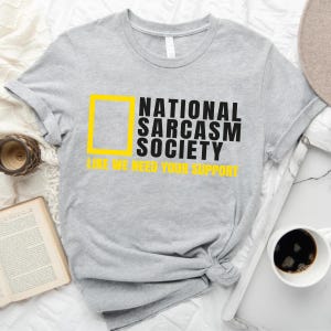 a gray t-shirt with a yellow graphic and text, along with a cup of coffee, a book, and a hat, all arranged on a bed.