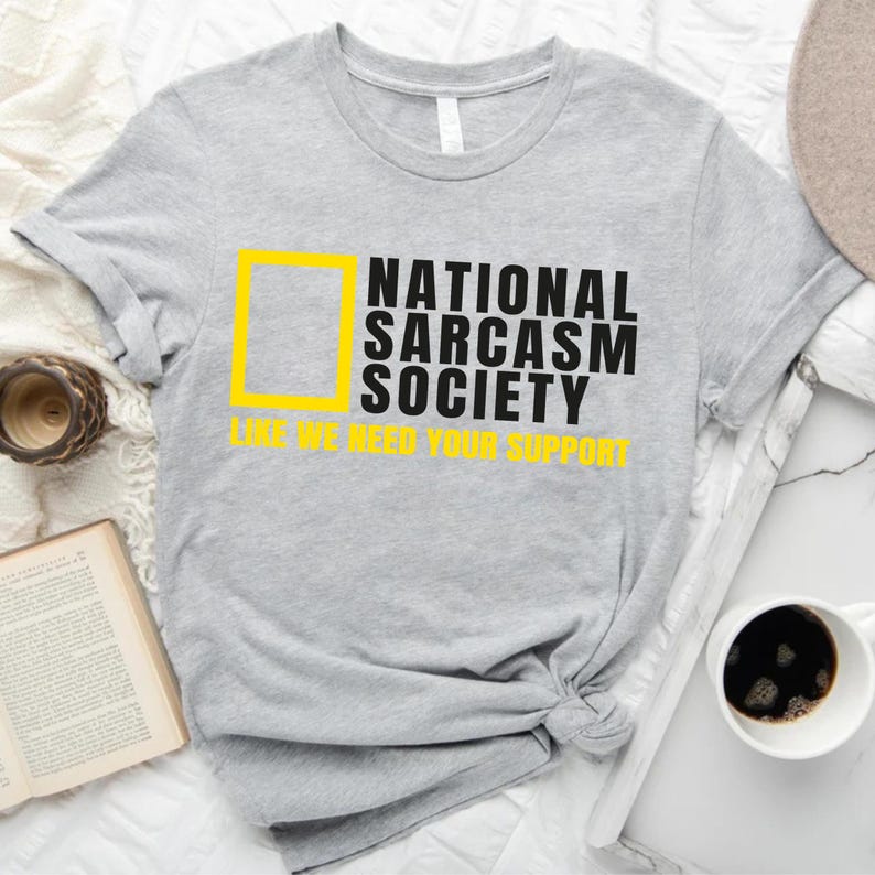 a gray t-shirt with a yellow graphic and text, along with a cup of coffee, a book, and a hat, all arranged on a bed.
