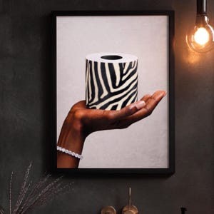 May include: Framed art print showing a roll of toilet paper with a black and white zebra stripe design. The print depicts a hand holding the toilet paper. A diamond bracelet is visible on the wrist. The art is in a bathroom.