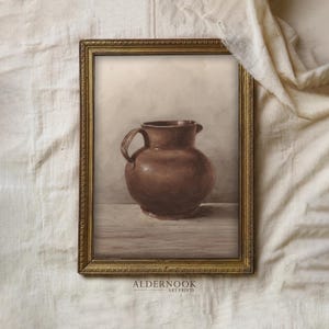 May include: A framed art print featuring a brown ceramic pitcher sitting on a flat surface. The pitcher has a rounded body, a wide rim, and a single handle on the left side. The artwork is enclosed in a decorative gold-toned frame with ornate detailing.