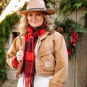 May include: A woman wearing a tan cowboy hat, a red and black plaid scarf, and a tan work jacket with a red lining. The jacket has a small logo patch on the front. The woman is smiling and standing in front of a wooden background with greenery.