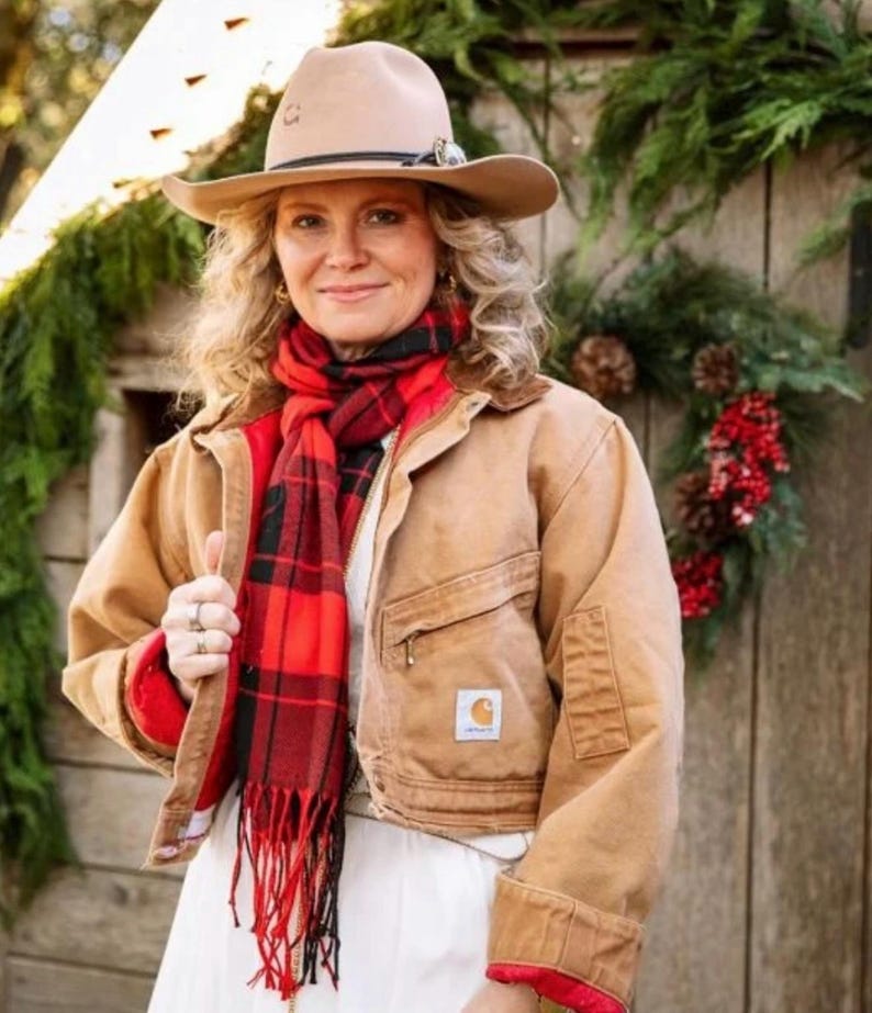 May include: A woman wearing a tan cowboy hat, a red and black plaid scarf, and a tan work jacket with a red lining. The jacket has a small logo patch on the front. The woman is smiling and standing in front of a wooden background with greenery.
