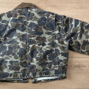 May include: A camouflage jacket with a brown corduroy collar. The jacket features a pattern of dark green, brown, and gray shapes. The jacket has two front pockets and a button closure. The sleeves have black cuffs.