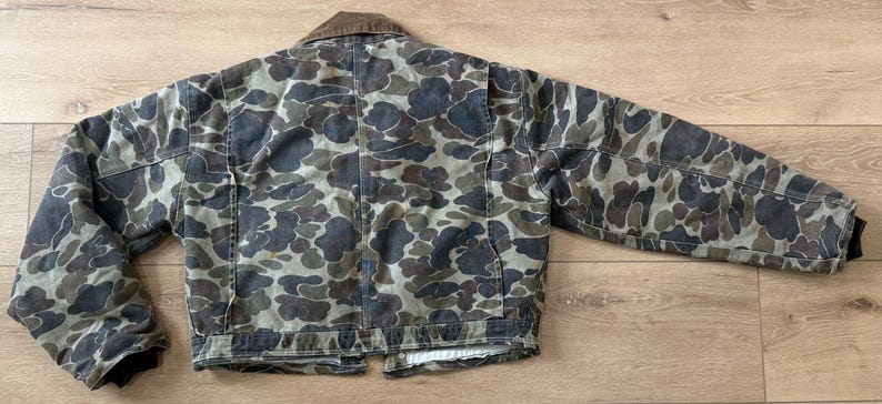 May include: A camouflage jacket with a brown corduroy collar. The jacket features a pattern of dark green, brown, and gray shapes. The jacket has two front pockets and a button closure. The sleeves have black cuffs.