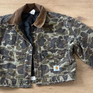 May include: A Carhartt camouflage jacket with a brown corduroy collar. The jacket has a button-up front, two chest pockets, and two lower pockets. The camouflage pattern features shades of brown, green, and gray.