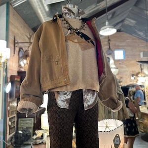 May include: A mannequin displays a tan work jacket, a cream-colored sweater with a southwestern design, and brown patterned pants. A white cowboy hat and a silver necklace complete the look. The outfit is displayed in a vintage shop.