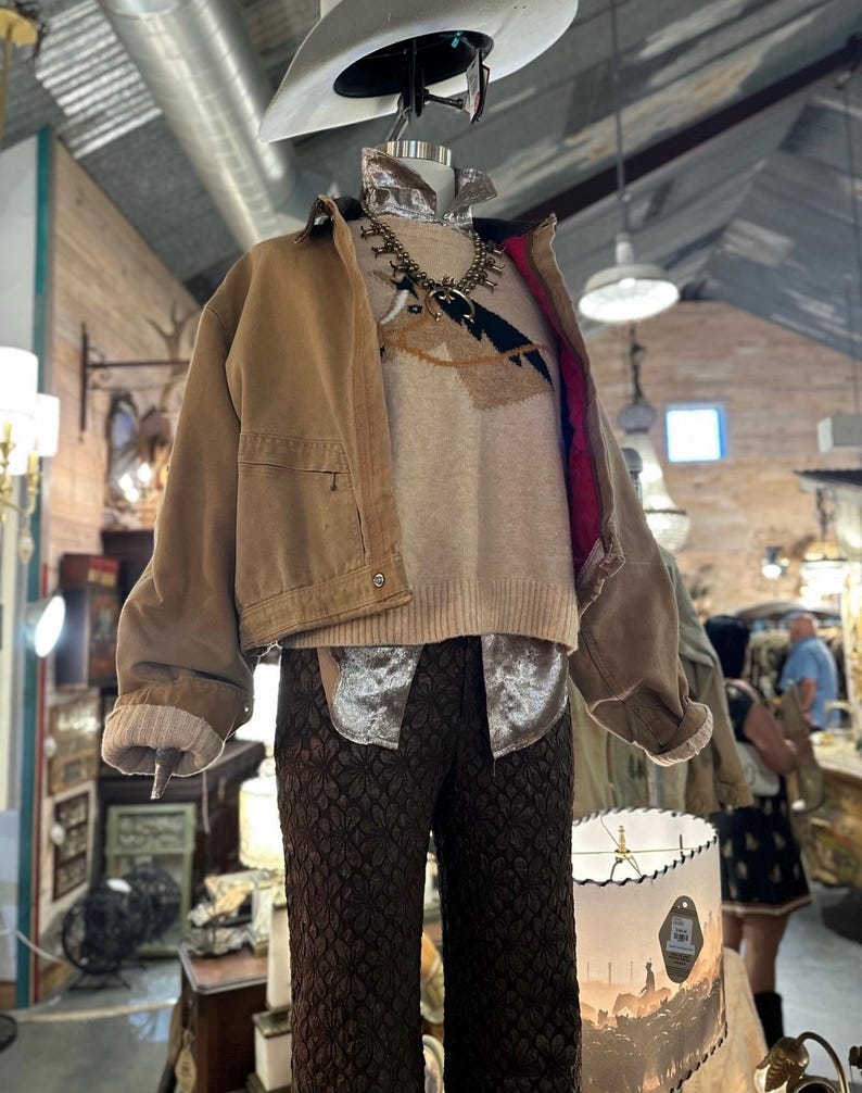 May include: A mannequin displays a tan work jacket, a cream-colored sweater with a southwestern design, and brown patterned pants. A white cowboy hat and a silver necklace complete the look. The outfit is displayed in a vintage shop.