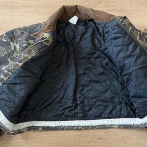 May include: Camouflage print jacket with a brown corduroy collar and a black quilted lining. The jacket is open, displaying the interior lining and the camouflage pattern on the exterior. The jacket is made of a durable material.