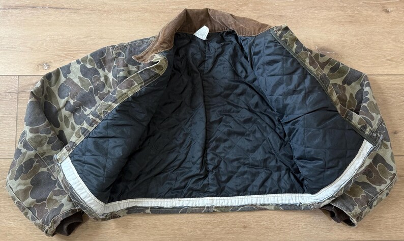 May include: Camouflage print jacket with a brown corduroy collar and a black quilted lining. The jacket is open, displaying the interior lining and the camouflage pattern on the exterior. The jacket is made of a durable material.