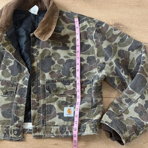 May include: Camouflage-patterned jacket with a brown corduroy collar. The jacket features multiple pockets, button closures, and the Carhartt logo. The jacket's length is approximately 20 inches.