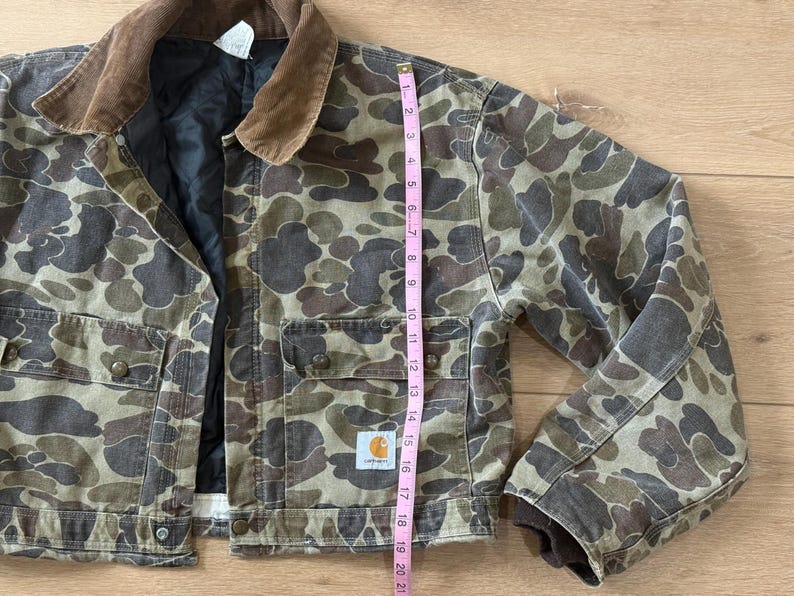 May include: Camouflage-patterned jacket with a brown corduroy collar. The jacket features multiple pockets, button closures, and the Carhartt logo. The jacket's length is approximately 20 inches.