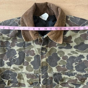 May include: Camouflage print jacket with a brown corduroy collar. The jacket has two front pockets and button closures. The jacket measures approximately 21 inches across the chest. The jacket is made of a durable material.