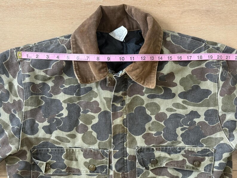 May include: Camouflage print jacket with a brown corduroy collar. The jacket has two front pockets and button closures. The jacket measures approximately 21 inches across the chest. The jacket is made of a durable material.