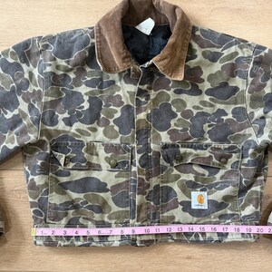 May include: A Carhartt duck camo jacket with a brown corduroy collar. The jacket has two chest pockets and two lower pockets. The jacket measures approximately 24 inches across the bottom. The Carhartt logo is visible on the lower pocket.