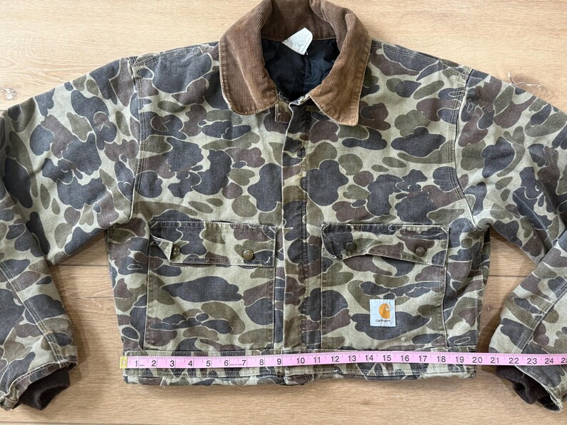 May include: A Carhartt duck camo jacket with a brown corduroy collar. The jacket has two chest pockets and two lower pockets. The jacket measures approximately 24 inches across the bottom. The Carhartt logo is visible on the lower pocket.