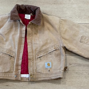 May include: A tan canvas jacket featuring a brown corduroy collar, a bright red quilted lining, and two front zippered pockets. The garment includes a small rectangular Carhartt brand patch on the lower left pocket and long sleeves with cuffs.
