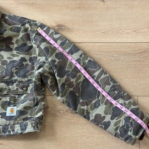 May include: Camouflage-patterned jacket with a brown corduroy collar. The jacket has two chest pockets and a Carhartt logo patch. The sleeve length is approximately 24 inches.
