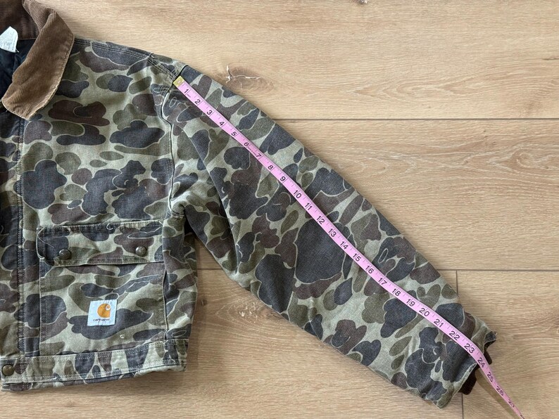 May include: Camouflage-patterned jacket with a brown corduroy collar. The jacket has two chest pockets and a Carhartt logo patch. The sleeve length is approximately 24 inches.