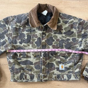 May include: Camouflage print jacket with a brown corduroy collar. The jacket has two chest pockets and two lower pockets. The jacket is approximately 28 inches wide. A small Carhartt logo is visible on the lower pocket.