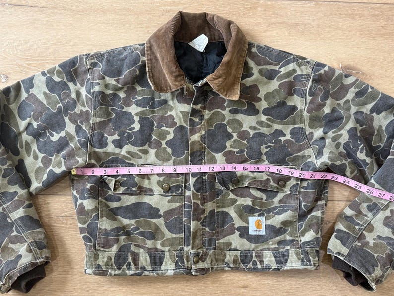 May include: Camouflage print jacket with a brown corduroy collar. The jacket has two chest pockets and two lower pockets. The jacket is approximately 28 inches wide. A small Carhartt logo is visible on the lower pocket.