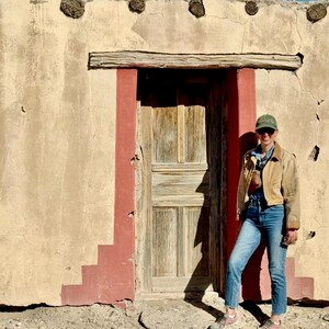 May include: A weathered wooden door with a red trim is set into a beige stucco wall. A person wearing a tan jacket, blue jeans, and a green cap stands in front of the door. The wall has decorative elements.