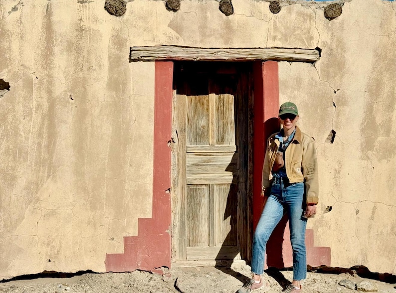May include: A weathered wooden door with a red trim is set into a beige stucco wall. A person wearing a tan jacket, blue jeans, and a green cap stands in front of the door. The wall has decorative elements.