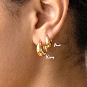 May include: Two gold hoop earrings with a smooth, polished finish. The smaller hoop is labeled 8mm and the larger hoop is labeled 10mm. These earrings feature a thick, rounded design and are worn in the earlobe, showcasing their size difference.