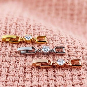 Puede incluir: Tres cierres de pulsera metálicos en oro, plata y oro rosa, cada uno con una gema redonda y transparente. Los cierres se muestran sobre un fondo de tela rosa texturizada, resaltando su diseño y variaciones de color.