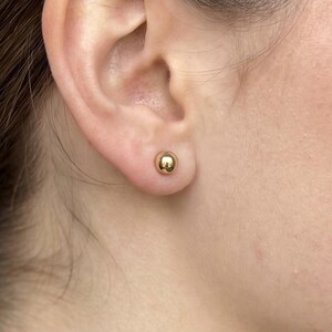 May include: A small, polished gold ball stud earring with a smooth, spherical surface. The earring is secured in the earlobe, featuring a reflective metallic finish that catches the light, creating a simple and classic circular shape.