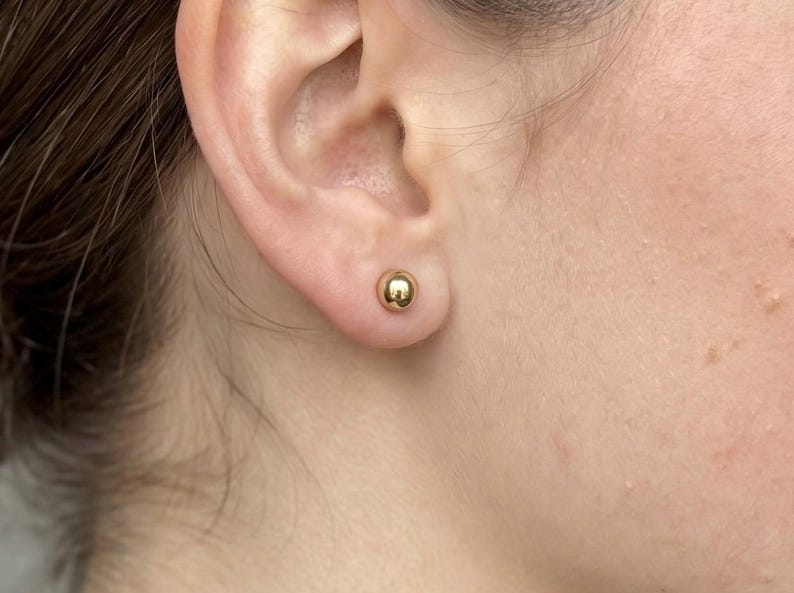 May include: A small, polished gold ball stud earring with a smooth, spherical surface. The earring is secured in the earlobe, featuring a reflective metallic finish that catches the light, creating a simple and classic circular shape.