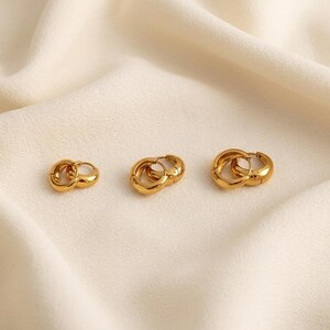 3 pairs of Gold-colored hoop earrings, 8mm, 10mm 12mm inner diameter size with their outer diameter being 15, 16, and 18mm respectively, are displayed on a soft ivory cream fabric. The image is a close-up, highlighting the jewelry's design and shine.