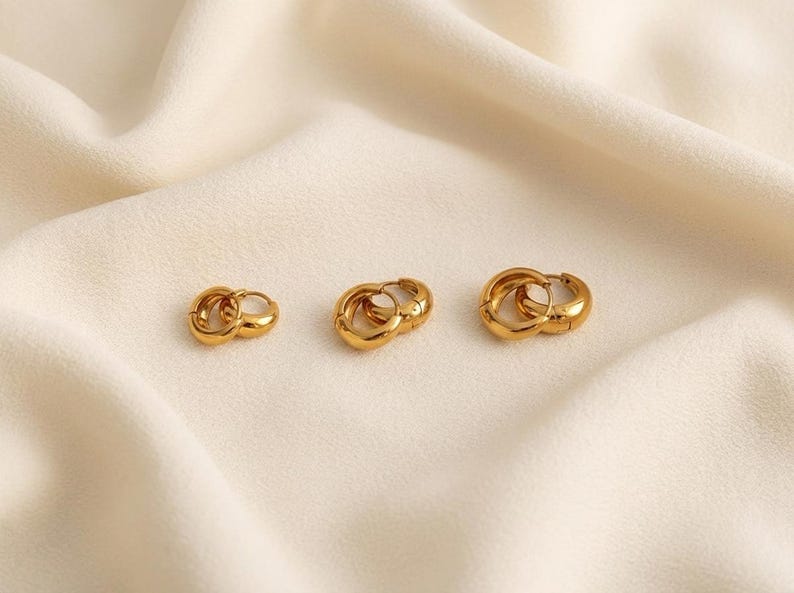 3 pairs of Gold-colored hoop earrings, 8mm, 10mm 12mm inner diameter size with their outer diameter being 15, 16, and 18mm respectively, are displayed on a soft ivory cream fabric. The image is a close-up, highlighting the jewelry's design and shine.