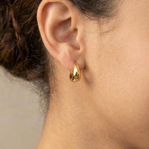Gold-colored hoop earrings, 12mm in size, are displayed on a right ear. The earrings have a smooth, polished finish and a classic, rounded shape. The image is a close-up, highlighting the jewelry's design and shine.