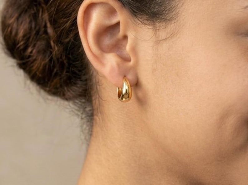 Gold-colored hoop earrings, 12mm in size, are displayed on a right ear. The earrings have a smooth, polished finish and a classic, rounded shape. The image is a close-up, highlighting the jewelry's design and shine.