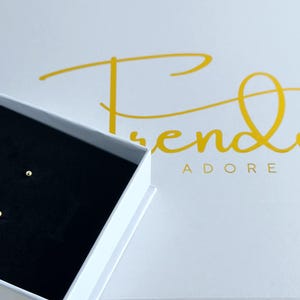 May include: A pair of small, round, gold-colored ball stud earrings resting inside a white jewelry box with a black velvet insert. The box lid features the gold-colored text Trendy ADORE in a cursive and block font combination.