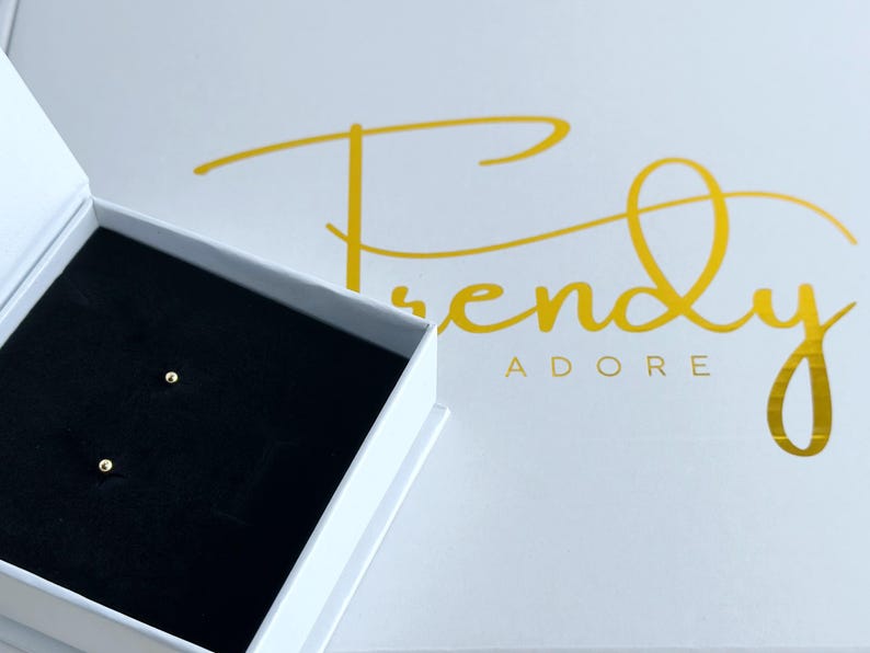 May include: A pair of small, round, gold-colored ball stud earrings resting inside a white jewelry box with a black velvet insert. The box lid features the gold-colored text Trendy ADORE in a cursive and block font combination.