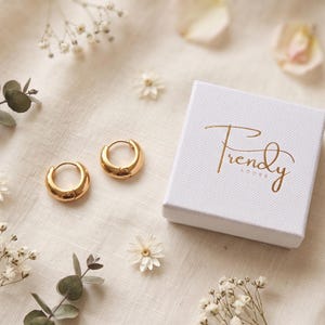 Gold-colored hoop earrings, 8mm in size, are displayed next to trendy adore jewelry box on a hand woven ivory cream fabric. The earrings have a smooth, polished finish and a classic, rounded shape. The image is a close-up, highlighting the jewelry's design and shine.