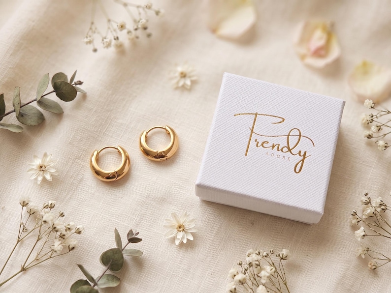 Gold-colored hoop earrings, 8mm in size, are displayed next to trendy adore jewelry box on a hand woven ivory cream fabric. The earrings have a smooth, polished finish and a classic, rounded shape. The image is a close-up, highlighting the jewelry's design and shine.