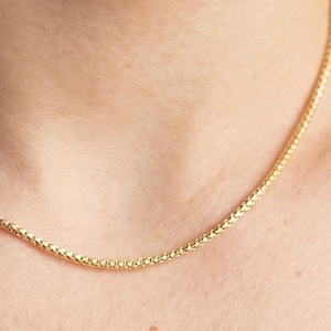 May include: A close-up of a gold-colored chain necklace. The chain has a woven design and is resting on a person's skin. The necklace is a simple, elegant piece of jewelry.