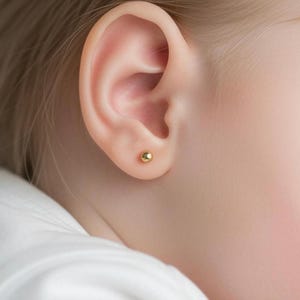 May include: A small, polished gold ball stud earring worn in the earlobe. The earring features a simple, spherical design with a smooth, reflective metallic surface that catches the light, providing a classic and understated appearance on the ear.