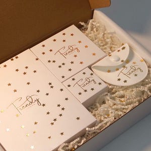 A white gift box containing a large rectangular box, three square boxes, and a semi-circular pouch, all decorated with gold star confetti and the gold script text "Trendy Adore". The items rest on a bed of white shredded paper filler.