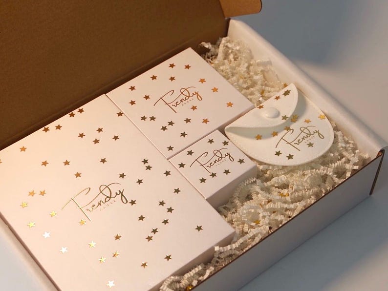 A white gift box containing a large rectangular box, three square boxes, and a semi-circular pouch, all decorated with gold star confetti and the gold script text "Trendy Adore". The items rest on a bed of white shredded paper filler.