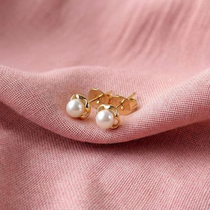 Könnte beinhalten: Ein Paar goldfarbene Ohrstecker mit Perlen in der Mitte, auf einem rosa Stoff. Die Ohrringe haben ein florales Design um die Perle. Der Hintergrund ist ein weiches, strukturiertes Rosa.
