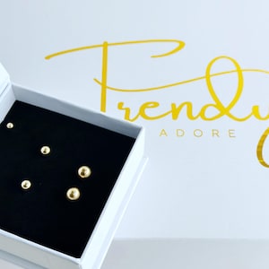 May include: A set of five gold-toned spherical stud earrings arranged on a black velvet insert inside a white square box. The box lid features the yellow text Trendy Adore. The earrings vary in size, presenting a polished, reflective metallic finish.